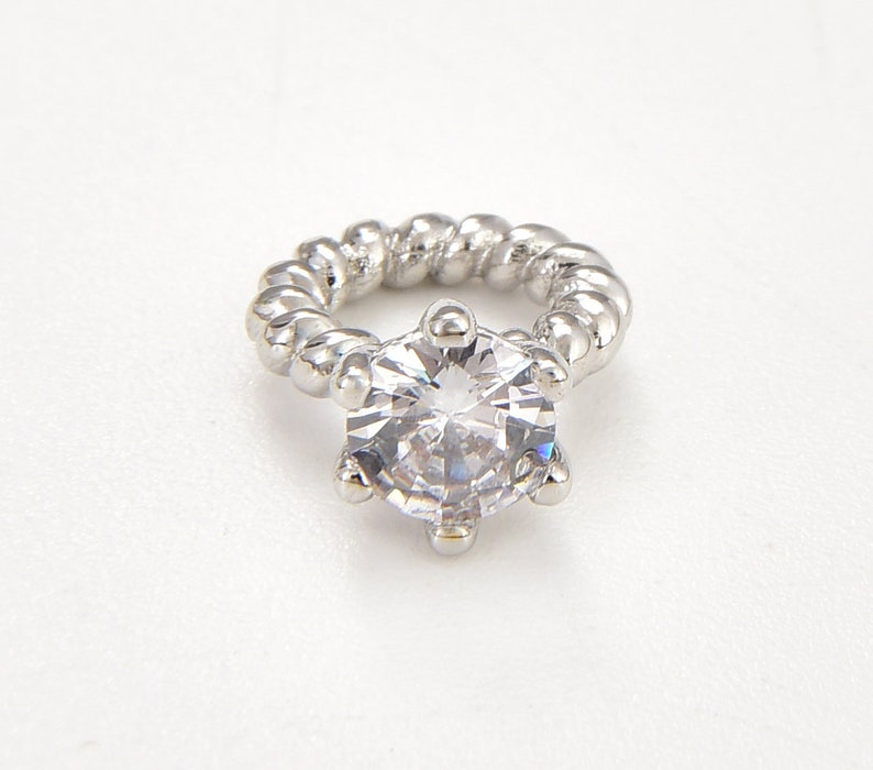 May include: A silver ring featuring a large, clear, faceted gemstone. The ring band has a twisted rope design. The gemstone is round with multiple facets, reflecting light. The ring is set against a white background.