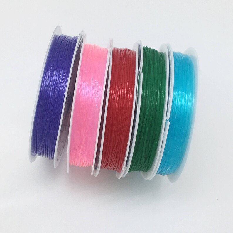 Puede incluir: Cinco carretes de cord&oacute;n el&aacute;stico en diferentes colores: morado, rosa, rojo, verde y azul. Los carretes est&aacute;n apilados uno encima del otro.