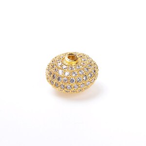 May include: A gold-toned bead with a round shape and a hole in the center. The bead is covered in small, clear rhinestones.
