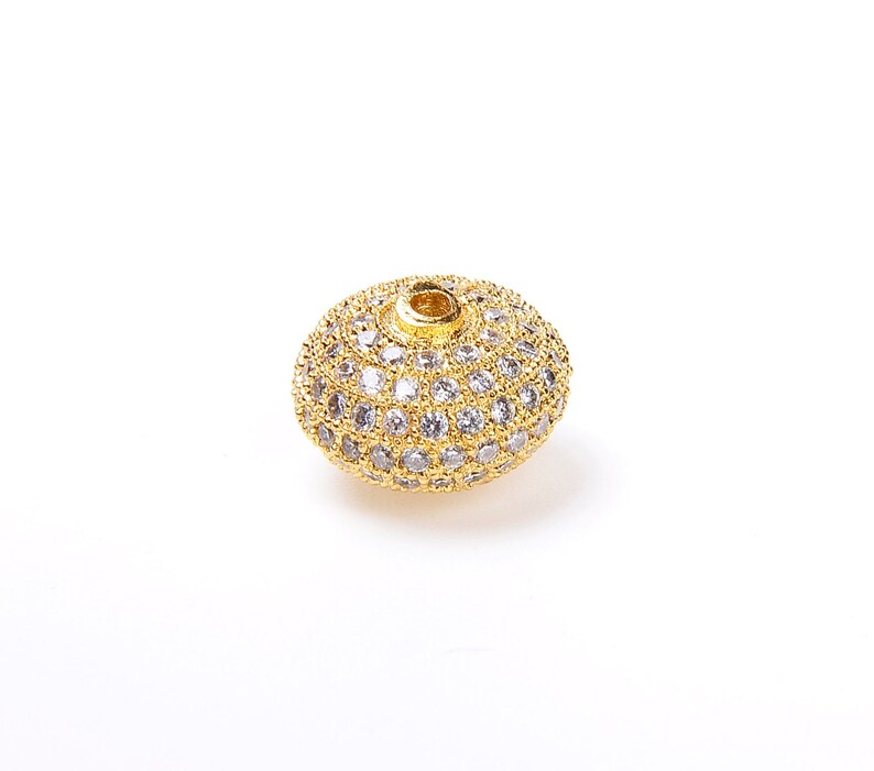 May include: A gold-toned bead with a round shape and a hole in the center. The bead is covered in small, clear rhinestones.