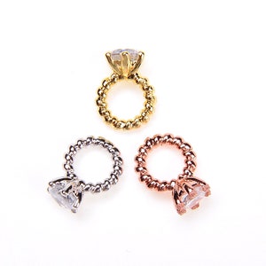 May include: Three engagement rings are shown, each with a clear, faceted stone and a twisted band. One is gold, one silver, and one rose gold. The rings are set against a white backdrop. These are likely jewellery.