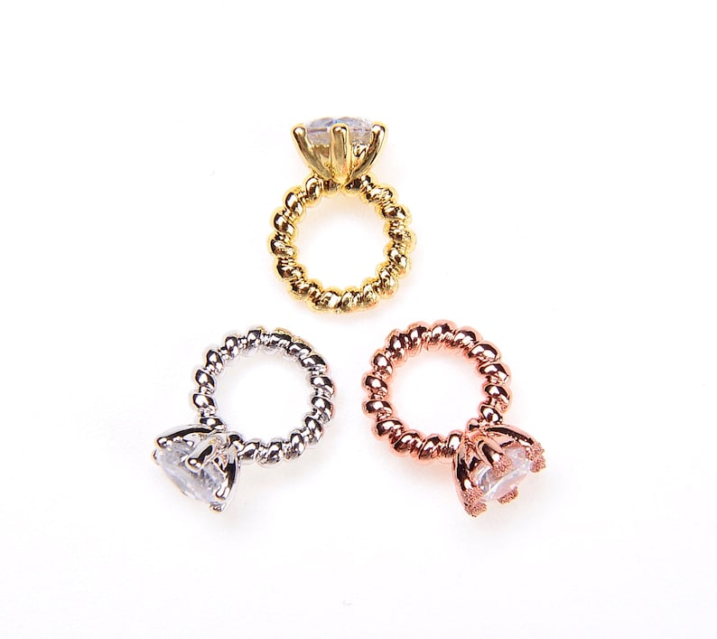 May include: Three engagement rings are shown, each with a clear, faceted stone and a twisted band. One is gold, one silver, and one rose gold. The rings are set against a white backdrop. These are likely jewellery.