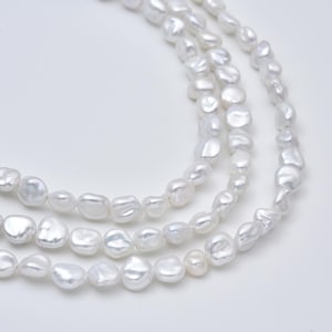 May include: A multi-strand necklace composed of numerous white, irregularly shaped baroque pearls. The pearls exhibit a high-luster, iridescent finish with a smooth, organic texture, arranged in three distinct, curved rows against a plain background.