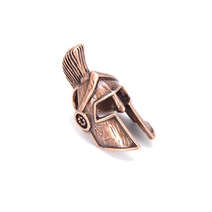 May include: A bronze-colored miniature Spartan helmet with a prominent crest and detailed facial features. The helmet is set against a white background, showcasing its intricate design and historical aesthetic. The helmet's color is a warm, metallic tone.
