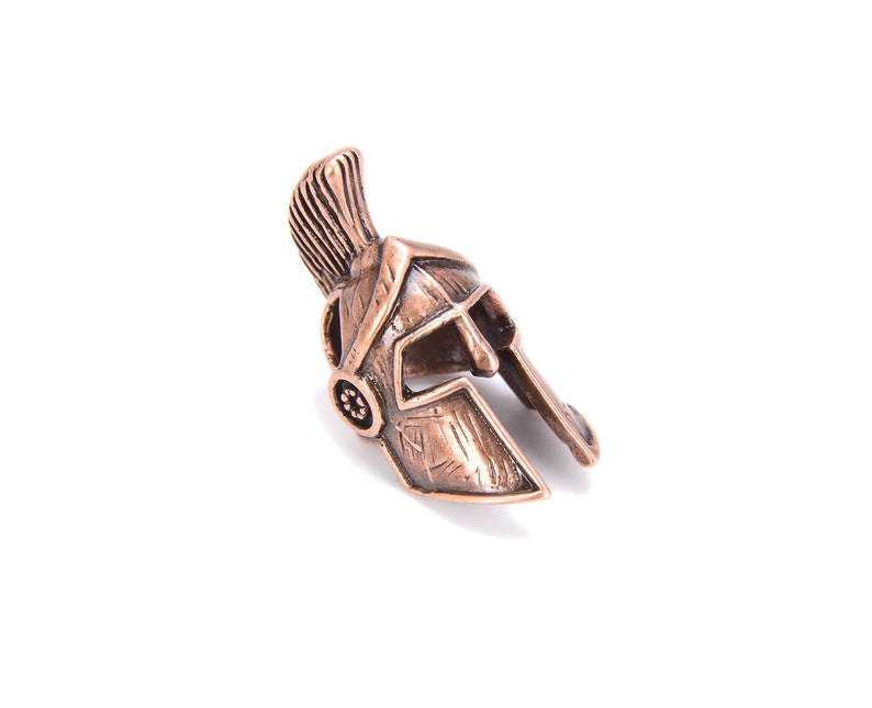 May include: A bronze-colored miniature Spartan helmet with a prominent crest and detailed facial features. The helmet is set against a white background, showcasing its intricate design and historical aesthetic. The helmet's color is a warm, metallic tone.
