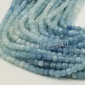 May include: A close-up of strands of aquamarine gemstone beads. The beads are a mix of light and dark blue, with a faceted, cube-like shape. The beads are strung together, creating a flowing, textured appearance. The image is well-lit, highlighting the color and detail of the gemstones.