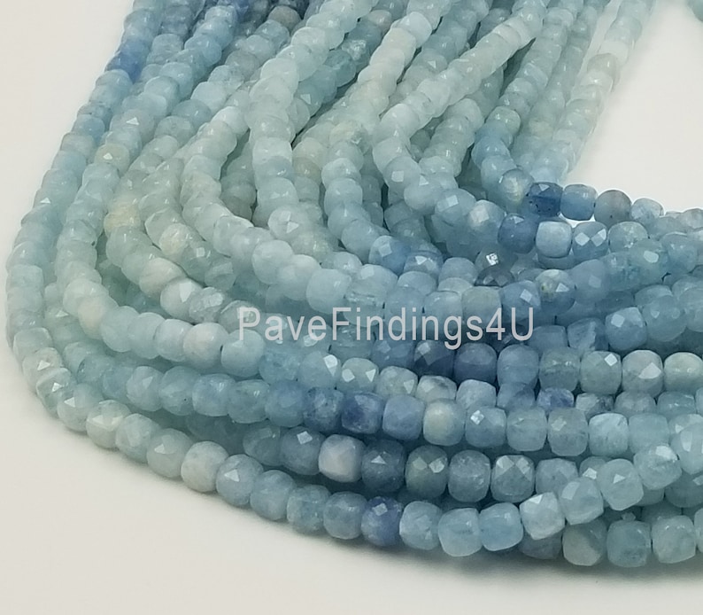 May include: A close-up of strands of aquamarine gemstone beads. The beads are a mix of light and dark blue, with a faceted, cube-like shape. The beads are strung together, creating a flowing, textured appearance. The image is well-lit, highlighting the color and detail of the gemstones.