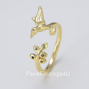 May include: Gold ring with a bird design and green gemstones on a branch. The bird is flying and has a clear gemstone for an eye.