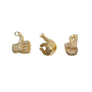 May include: Three gold-tone charms in the shape of hands. The first charm is a thumbs up, the second charm is a peace sign, and the third charm is an OK sign. All charms are covered in clear rhinestones.