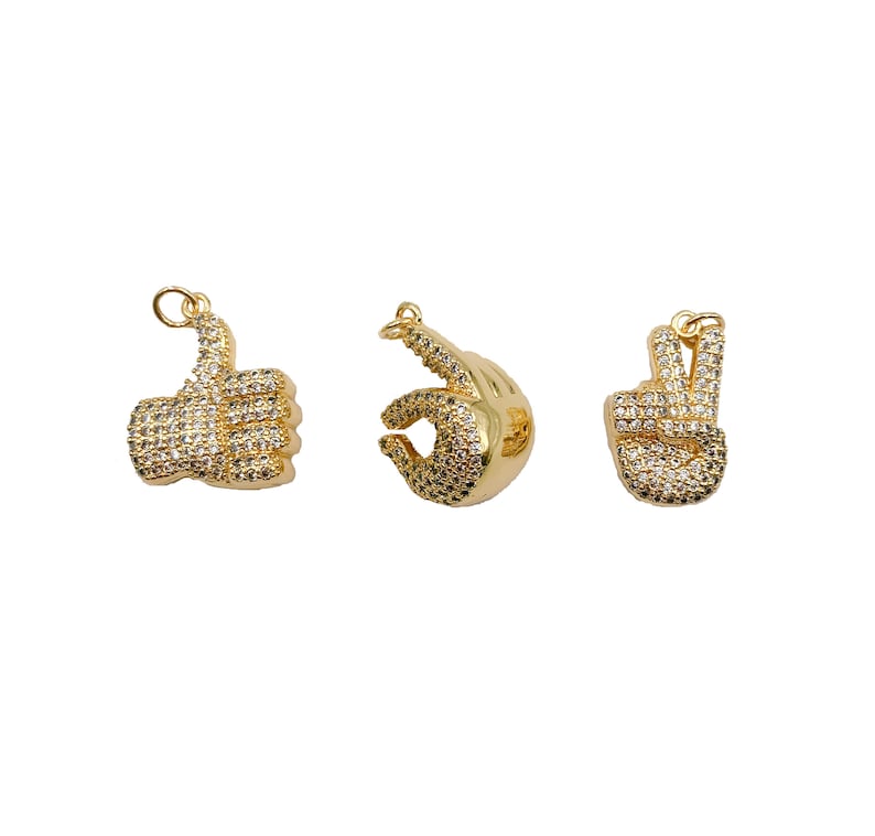 May include: Three gold-tone charms in the shape of hands. The first charm is a thumbs up, the second charm is a peace sign, and the third charm is an OK sign. All charms are covered in clear rhinestones.
