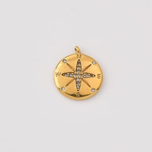 May include: A gold-colored compass pendant with a loop at the top for a chain. The compass rose is detailed with small, sparkling stones. The cardinal directions are marked with the letters N, S, E, and W.