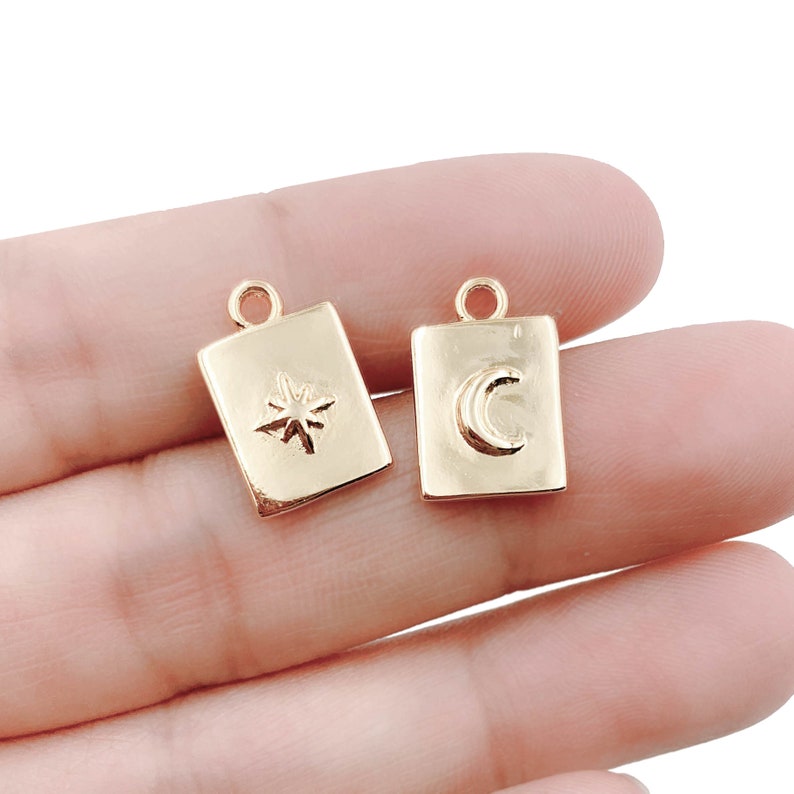 May include: Two gold-toned metal charms in the shape of a square. One charm features a star design and the other charm features a crescent moon design.