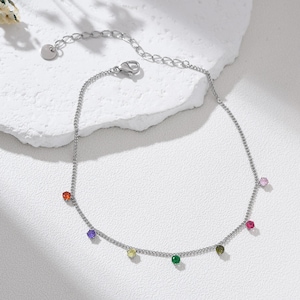 May include: A silver-colored anklet with a delicate chain design. The anklet features a series of colorful, round gemstones in red, purple, yellow, green, pink, and olive green. The anklet is displayed on a white surface.