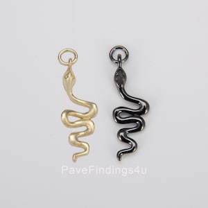 Pode incluir: Dois pingentes de metal em forma de cobra, um em tom dourado e outro em tom preto, cada um com um pequeno anel circular no topo para fixação. O texto PaveFindings4u está impresso em cinza claro na parte inferior central da imagem.
