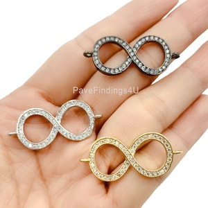 May include: Three infinity charm connectors in silver, gold, and black. Each charm is encrusted with small, clear crystals. The charms are held in a hand, showcasing the different colour options available.