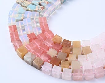 Peut inclure: Gros plan de plusieurs brins de perles cubiques de différentes couleurs, dont rose, pêche, bleu et beige. Les perles sont enfilées ensemble, créant une forme incurvée. Les perles semblent être en pierre ou en verre.