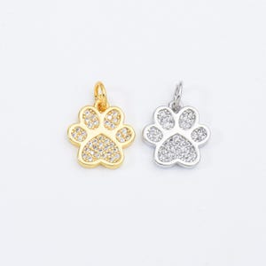 May include: Two paw print charms, one gold-toned and one silver-toned, each encrusted with small, clear crystals. The charms have a loop at the top for attaching to a necklace or bracelet. The background is plain white.