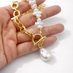 May include: A gold-toned necklace featuring a toggle clasp and a chain with irregular links. The necklace has a strand of white, irregularly shaped pearls and a large, pear-shaped pearl pendant.