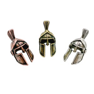 May include: Three miniature Spartan helmet pendants in copper, brass, and silver tones. Each helmet features a detailed crest and face guard, set against a white background. These pendants are likely for jewelry or crafting.