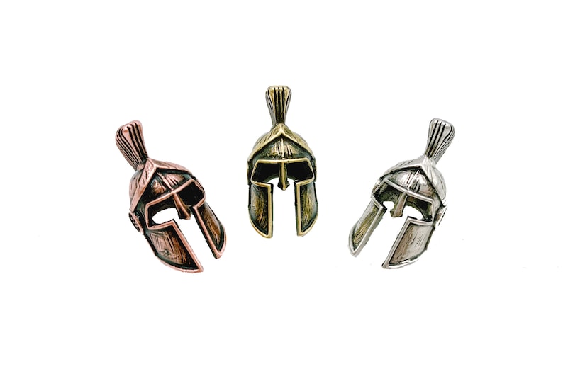 May include: Three miniature Spartan helmet pendants in copper, brass, and silver tones. Each helmet features a detailed crest and face guard, set against a white background. These pendants are likely for jewelry or crafting.