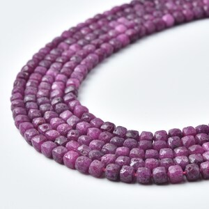 May include: Close-up of strands of faceted, deep magenta-colored gemstone beads. The beads are square-shaped and strung together, creating a textured, organic look. The background is plain white, highlighting the rich color of the gemstones.