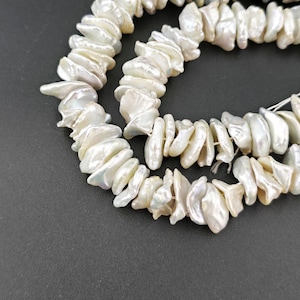 Puede incluir: Primer plano de hilos de perlas de agua dulce irregulares, blanquecinas. Las perlas tienen una forma natural y orgánica y un brillo sutil. El fondo es gris oscuro, proporcionando contraste. Adecuado para la creación de joyas.