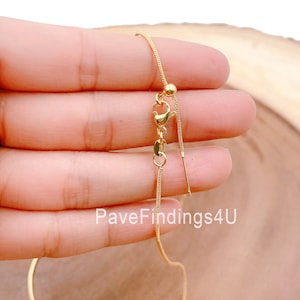 May include: A gold-toned adjustable chain necklace with a sliding clasp and a small bead at the end of the chain. The chain is shown on a hand against a wooden background.