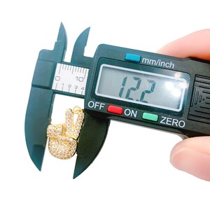 May include: A gold peace sign charm with clear rhinestones is measured with a digital caliper. The caliper reads 12.2 mm.