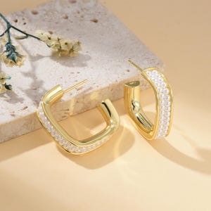 May include: A pair of gold-coloured, square hoop earrings. Each earring is lined with small, white pearls along the outer edge. The earrings are displayed on a textured, light-coloured surface with a sprig of dried flowers.