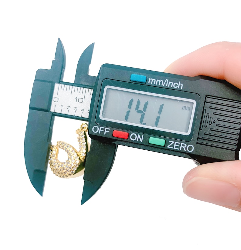 May include: A black digital caliper measuring a gold-colored pendant with clear stones. The caliper reads 14.1 mm.
