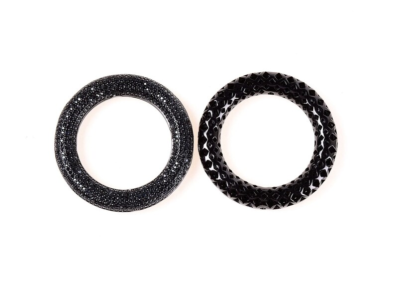 May include: Two black bangles. One is covered in small, sparkling black stones. The other has a geometric, openwork design. Both are circular and sit against a white background.