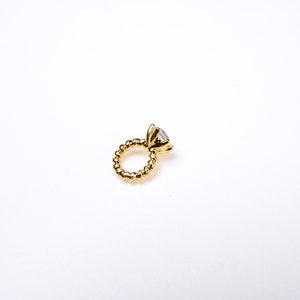 May include: A gold ring with a large, clear gemstone. The ring's band has a textured, beaded design. The ring is set against a plain white background, highlighting its details.