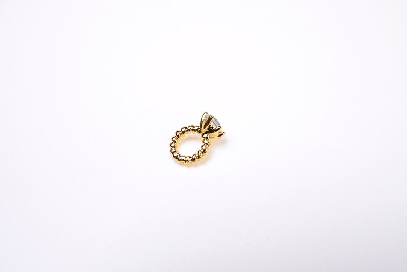 May include: A gold ring with a large, clear gemstone. The ring's band has a textured, beaded design. The ring is set against a plain white background, highlighting its details.
