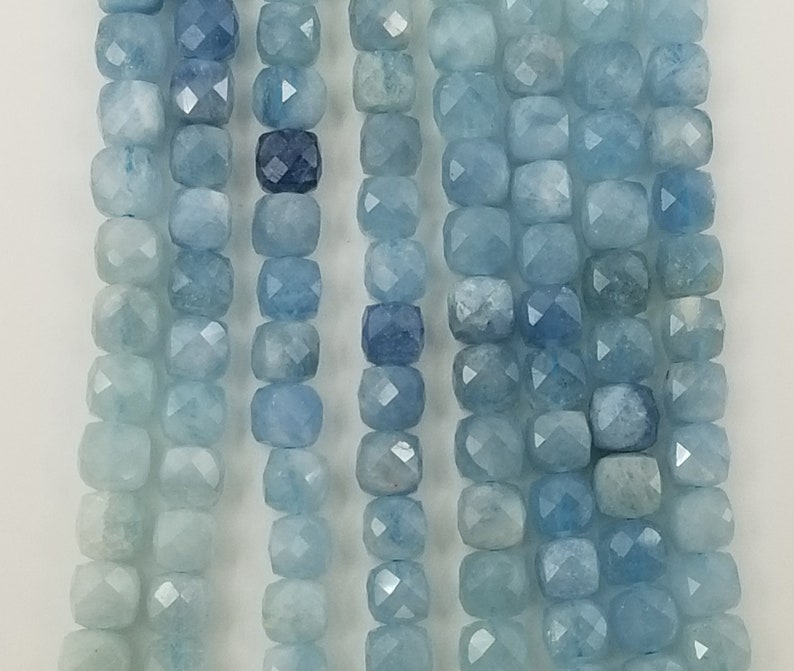 May include: Strands of aquamarine gemstone beads in varying shades of blue. The beads are square-cut with faceted surfaces, creating a sparkling effect. The color palette ranges from pale to deep blue.