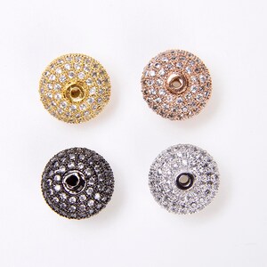 May include: Four round, flat, decorative beads with a hole in the center. The beads are covered in small, clear rhinestones. The beads are in four different colors: gold, rose gold, black, and silver.
