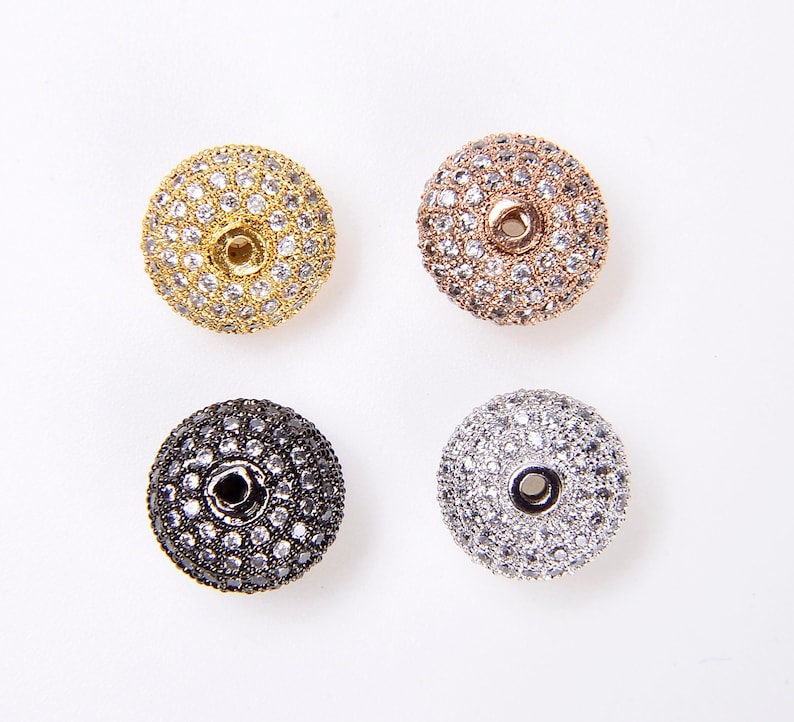 May include: Four round, flat, decorative beads with a hole in the center. The beads are covered in small, clear rhinestones. The beads are in four different colors: gold, rose gold, black, and silver.