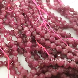 May include: A strand of ruby red faceted beads, strung on pink thread. The beads are small and round, with a slightly irregular shape. The beads are all approximately the same size.