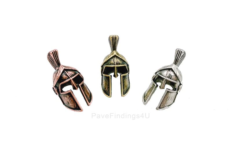 May include: Three miniature Spartan helmet charms in bronze, silver, and copper tones. Each helmet features a detailed crest and face guard, perfect for jewelry making or crafting projects. The charms are displayed against a white background.