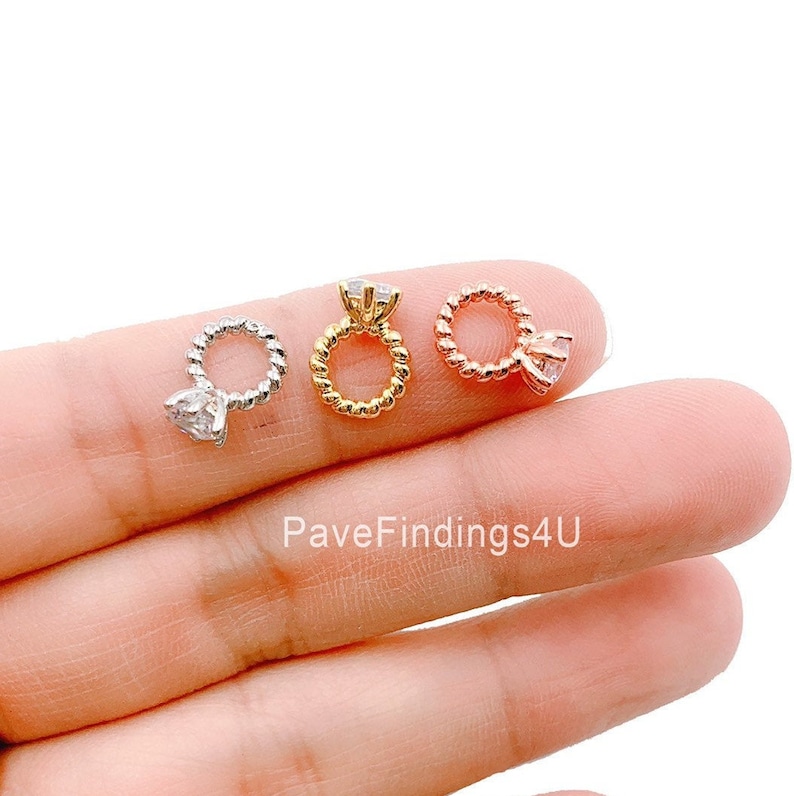 May include: Three small ring-shaped charms in silver, gold, and rose gold. Each charm features a twisted band and a clear, faceted gemstone. The charms are displayed on a light background.