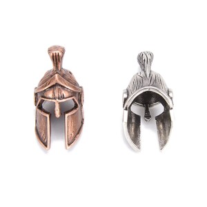 May include: Two miniature Spartan helmet pendants. One is bronze-colored, the other is silver-colored. Both have detailed designs, including a crest and face guard. The helmets are displayed against a white background.