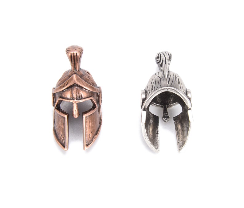 May include: Two miniature Spartan helmet pendants. One is bronze-colored, the other is silver-colored. Both have detailed designs, including a crest and face guard. The helmets are displayed against a white background.