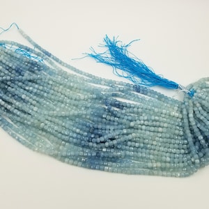 May include: Strands of aquamarine gemstone beads in varying shades of blue. The beads are small, square-cut, and strung together, with a bright blue tassel at the end. The beads are displayed on a white surface.