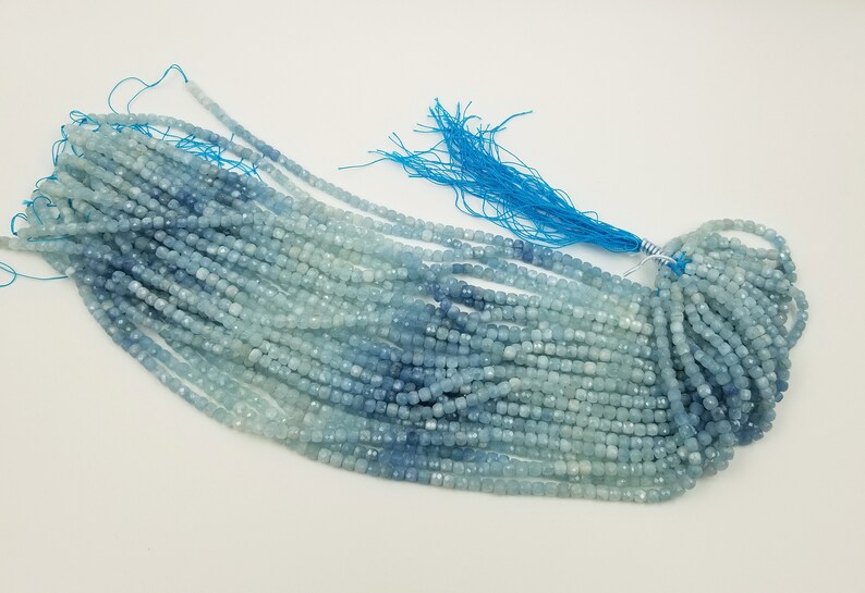 May include: Strands of aquamarine gemstone beads in varying shades of blue. The beads are small, square-cut, and strung together, with a bright blue tassel at the end. The beads are displayed on a white surface.
