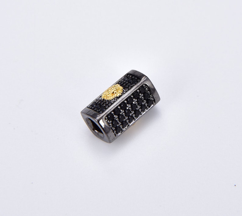 May include: Black and gold rectangular bead with a lion head design. The bead is covered in small black stones.