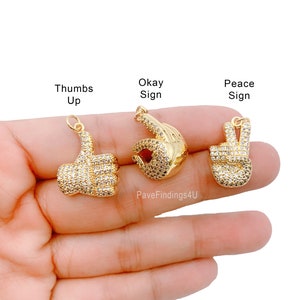 May include: Three gold-tone charms with clear crystal accents. The charms are shaped like a thumbs up, an okay sign, and a peace sign.