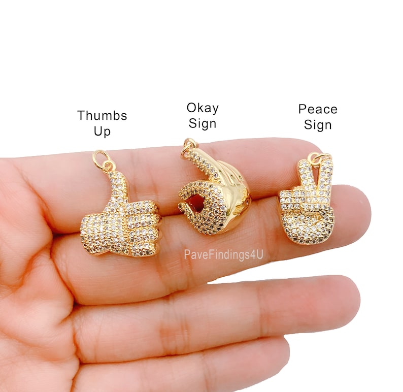 May include: Three gold-tone charms with clear crystal accents. The charms are shaped like a thumbs up, an okay sign, and a peace sign.