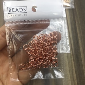 May include: A clear plastic bag filled with a large quantity of small, rose gold jump rings. The bag has a white label with the text "BEADS CREATION4U".