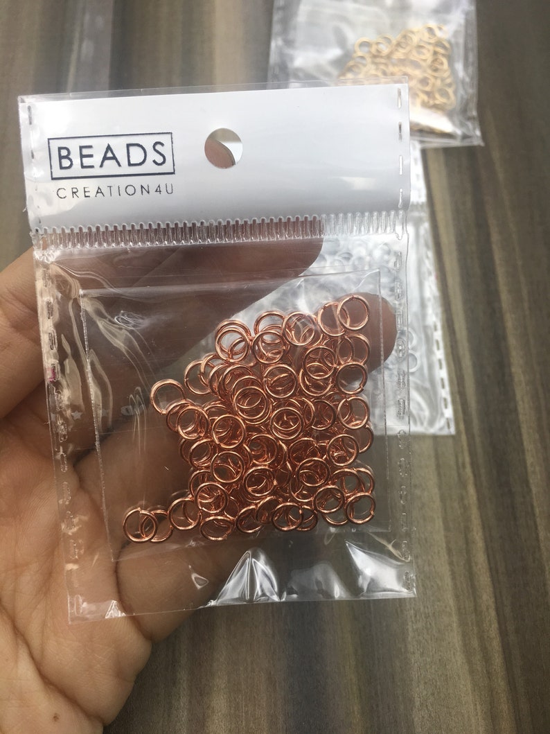 May include: A clear plastic bag filled with a large quantity of small, rose gold jump rings. The bag has a white label with the text "BEADS CREATION4U".