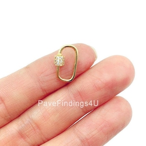 May include: Gold-toned metal clasp with a small, square-shaped, clear crystal-encrusted charm. The clasp is open and is being held by a hand.