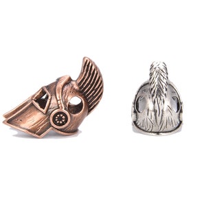 May include: Two metal helmet beads. One is bronze-colored with a detailed design, including a crest and circular accents. The other is silver-colored, also with a crest and detailed features. Both are on a white background.